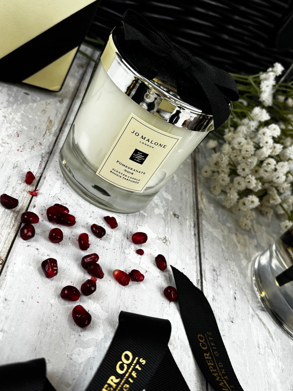 A Jo Malone London Pomegranate scented candle in a clear glass jar with a cream-colored wax, a black ribbon bow, and a gold label, styled on a white wooden surface with scattered pomegranate seeds, black ribbons, and white flowers.