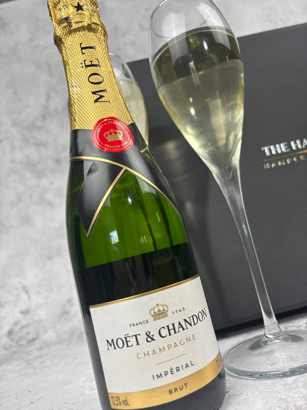 A bottle of Moët & Chandon Impérial Brut champagne next to a filled champagne flute, with a dark branded box in the background on a light stone surface.