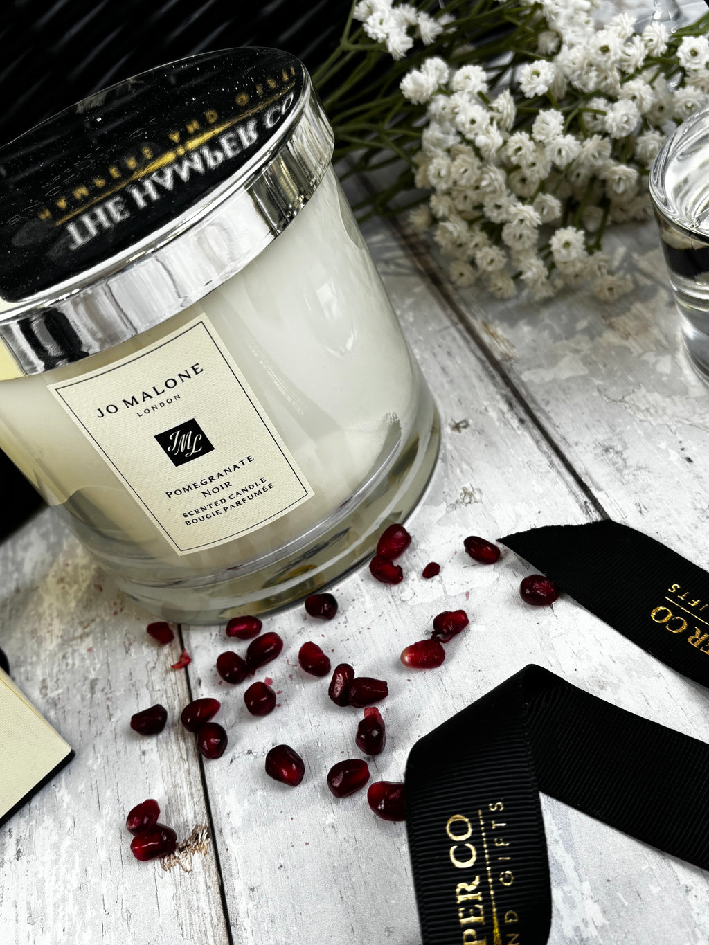 A Jo Malone London candle in a clear glass jar with a black lid and cream label, styled on a white wooden surface with scattered pomegranate seeds, white flowers, and black ribbon accents.