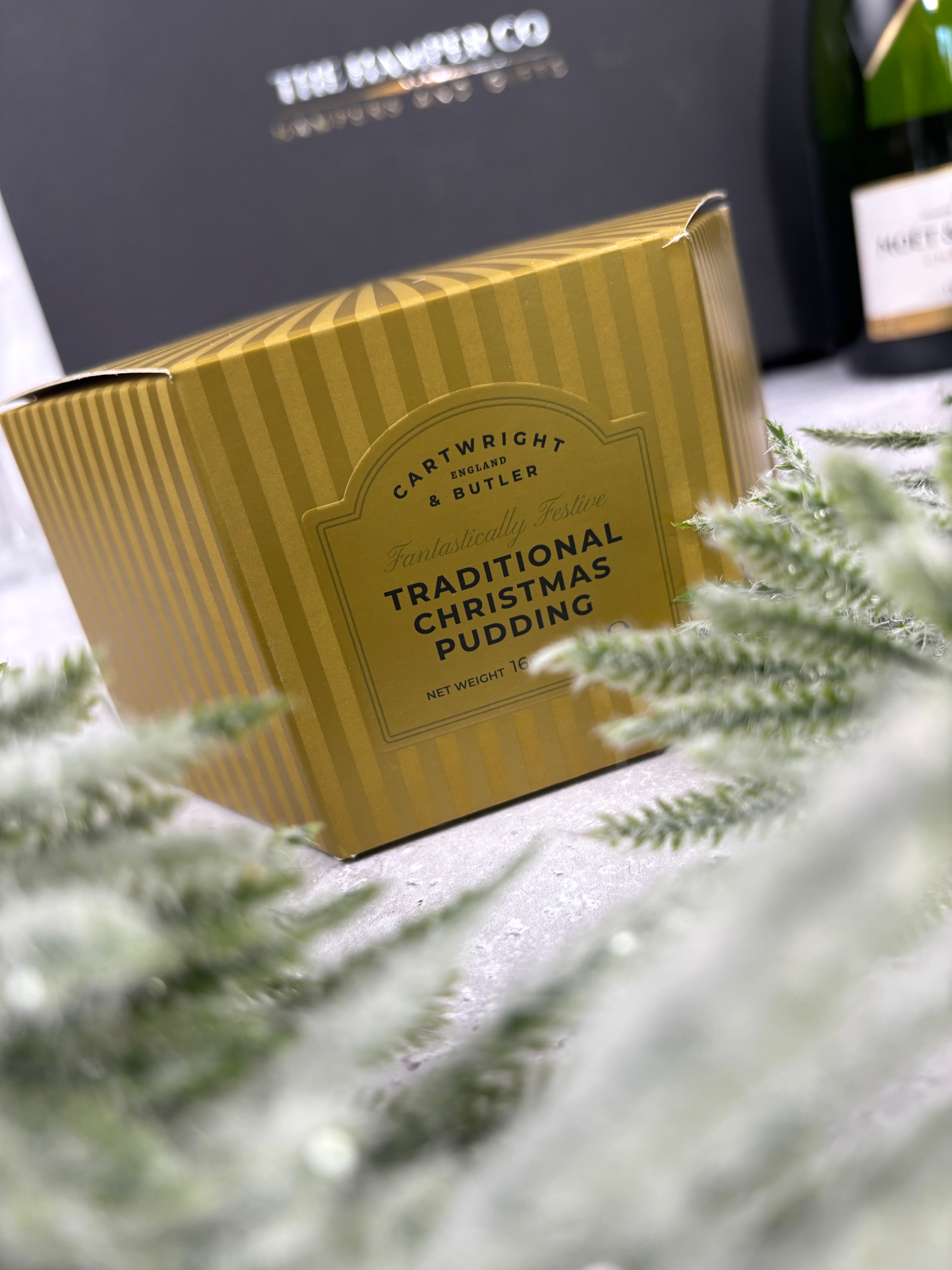 A gold striped box of Cartwright & Butler Traditional Christmas Pudding, styled with blurred green pine branches in the foreground on a light gray surface.