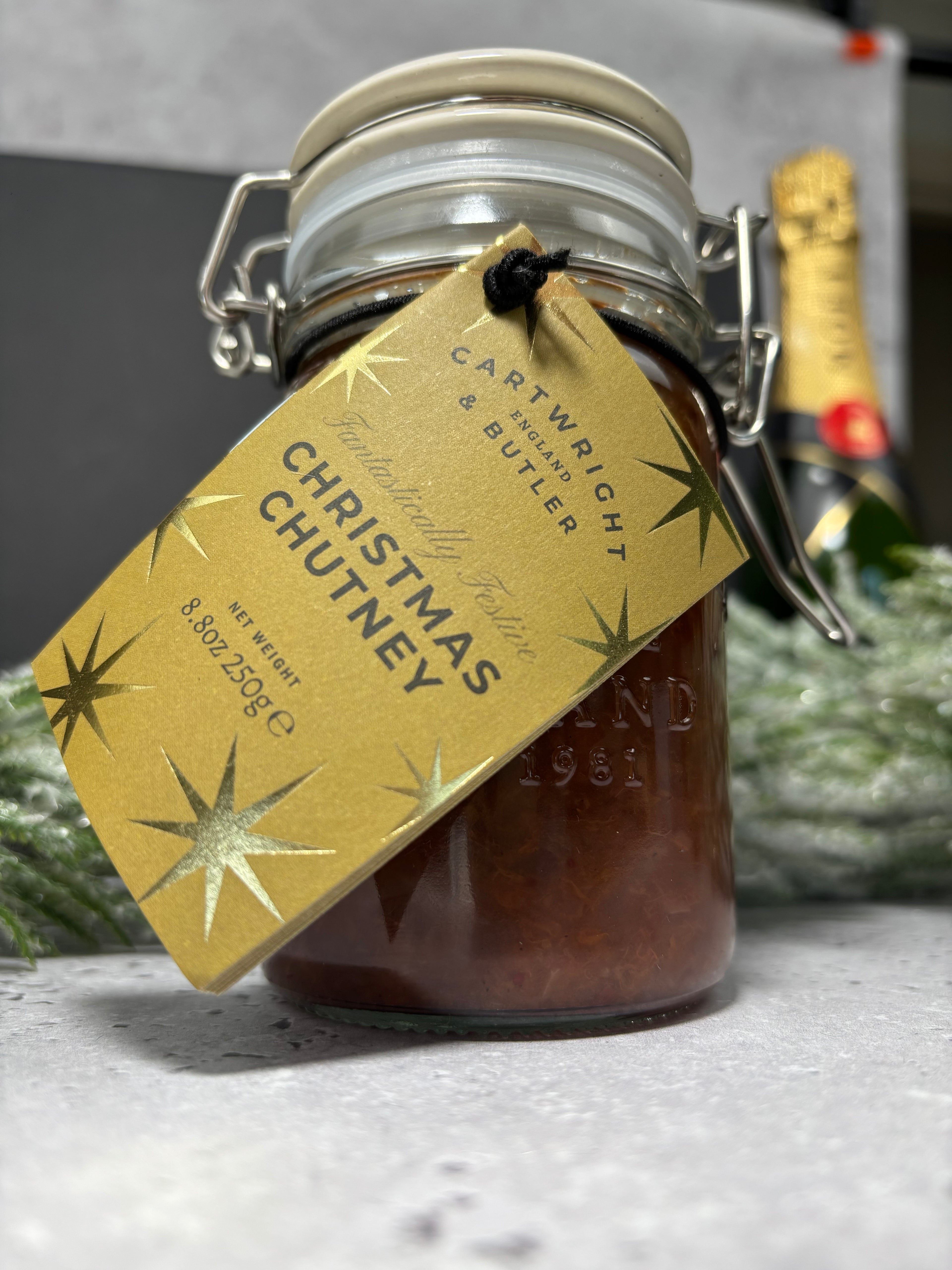A glass clamp-lid jar filled with Christmas chutney, with a gold paper tag reading 'Cartwright & Butler Christmas Chutney' and gold starburst graphics. A champagne bottle and greenery are blurred in the background on a light gray surface.