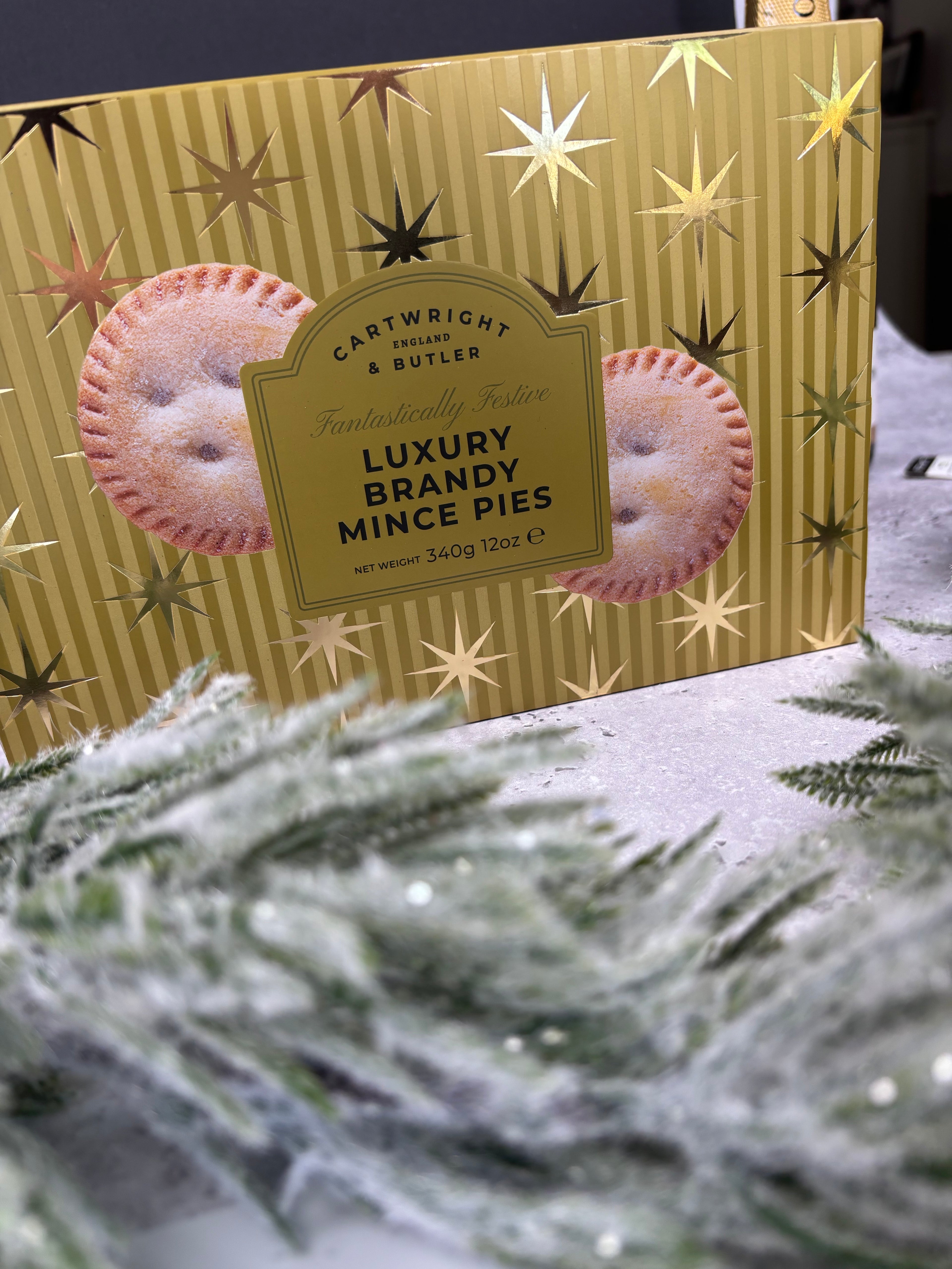 A box of Cartwright & Butler Luxury Brandy Mince Pies, featuring a gold striped background with metallic star patterns and illustrations of two mince pies. The box is styled on a snowy surface with blurred green pine branches in the foreground.