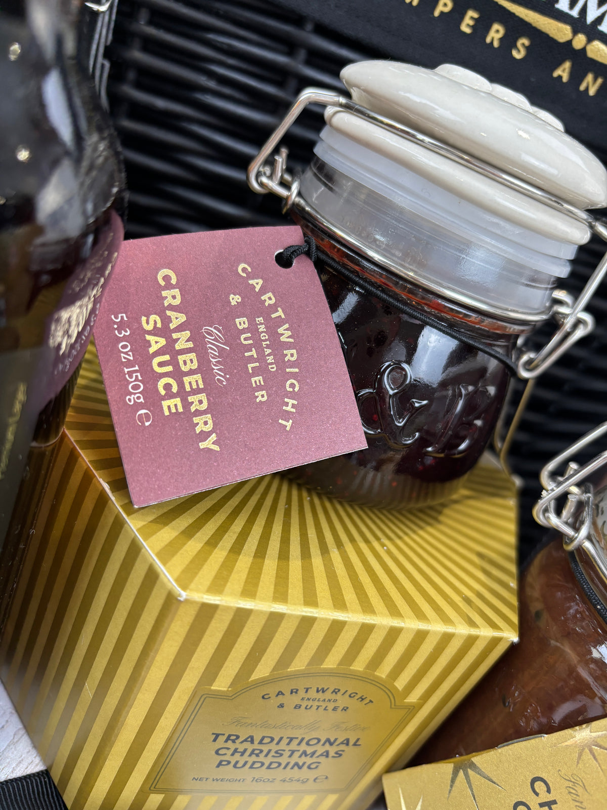 A close-up of a glass jar with a white lid and a pink hanging tag reading 'Cartwright & Butler Cranberry Sauce,' placed on top of a yellow striped box labeled 'Traditional Christmas Pudding.'.