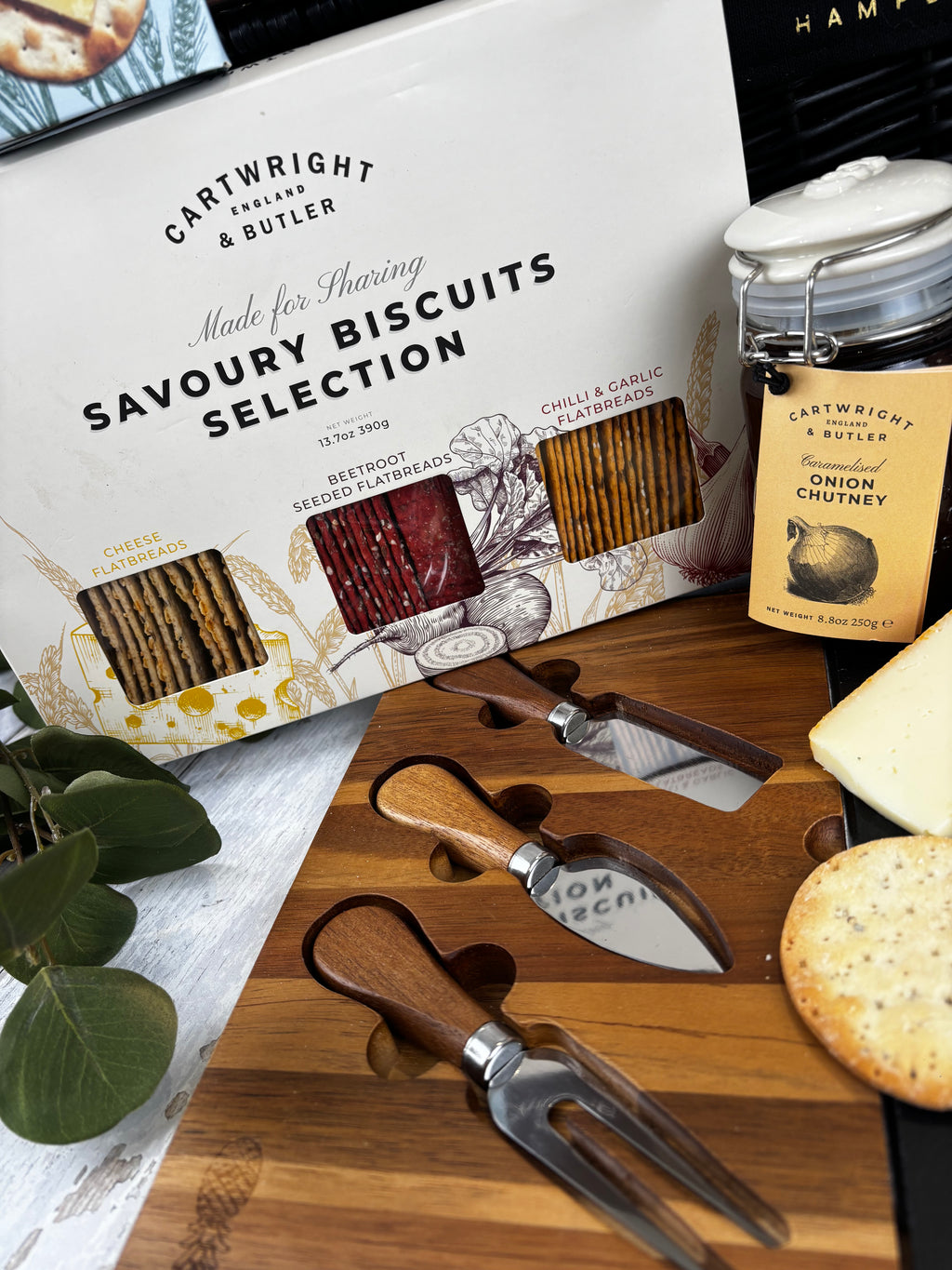 Cartright Selection Savoury Biscuit Set displayed on a wooden board with crackers, cheese, a glass jar, and green leaves. The set includes three knives and two forks in a gift box with a wooden stand.