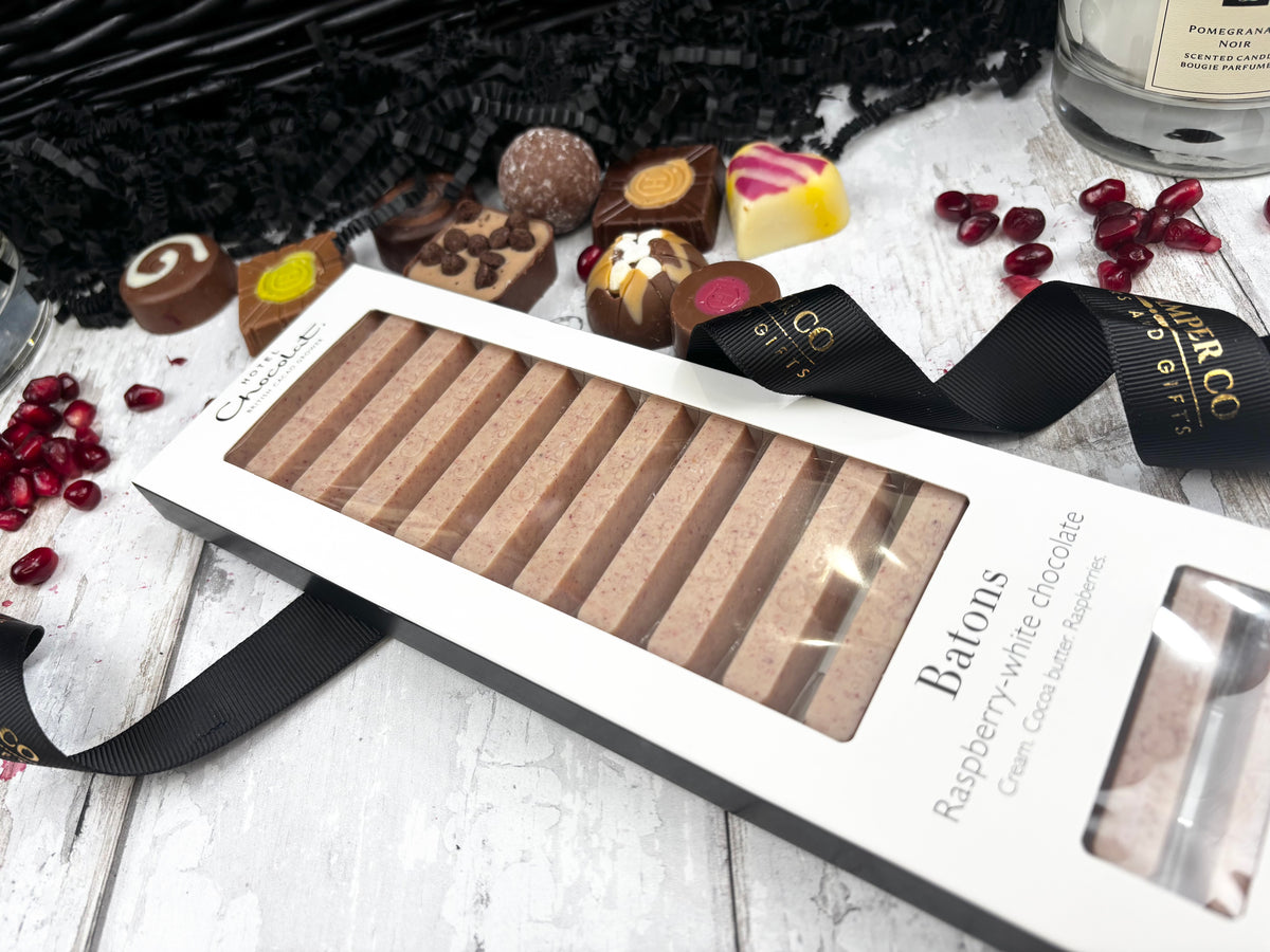 A boxed set of chocolate batons arranged on a white wooden surface with decorative chocolates, pomegranate seeds, black tinsel, and a black ribbon. The package is labeled 'Creme Brulee' and 'Batons Raspberry white chocolate'.