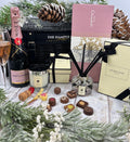 A luxury gift hamper arrangement featuring a Moët & Chandon Rosé bottle, a Hamper Co basket, Jo Malone candle and reed diffuser, a pink chocolate box, and assorted chocolates, styled with pine branches and pinecones on a rustic white wooden surface.