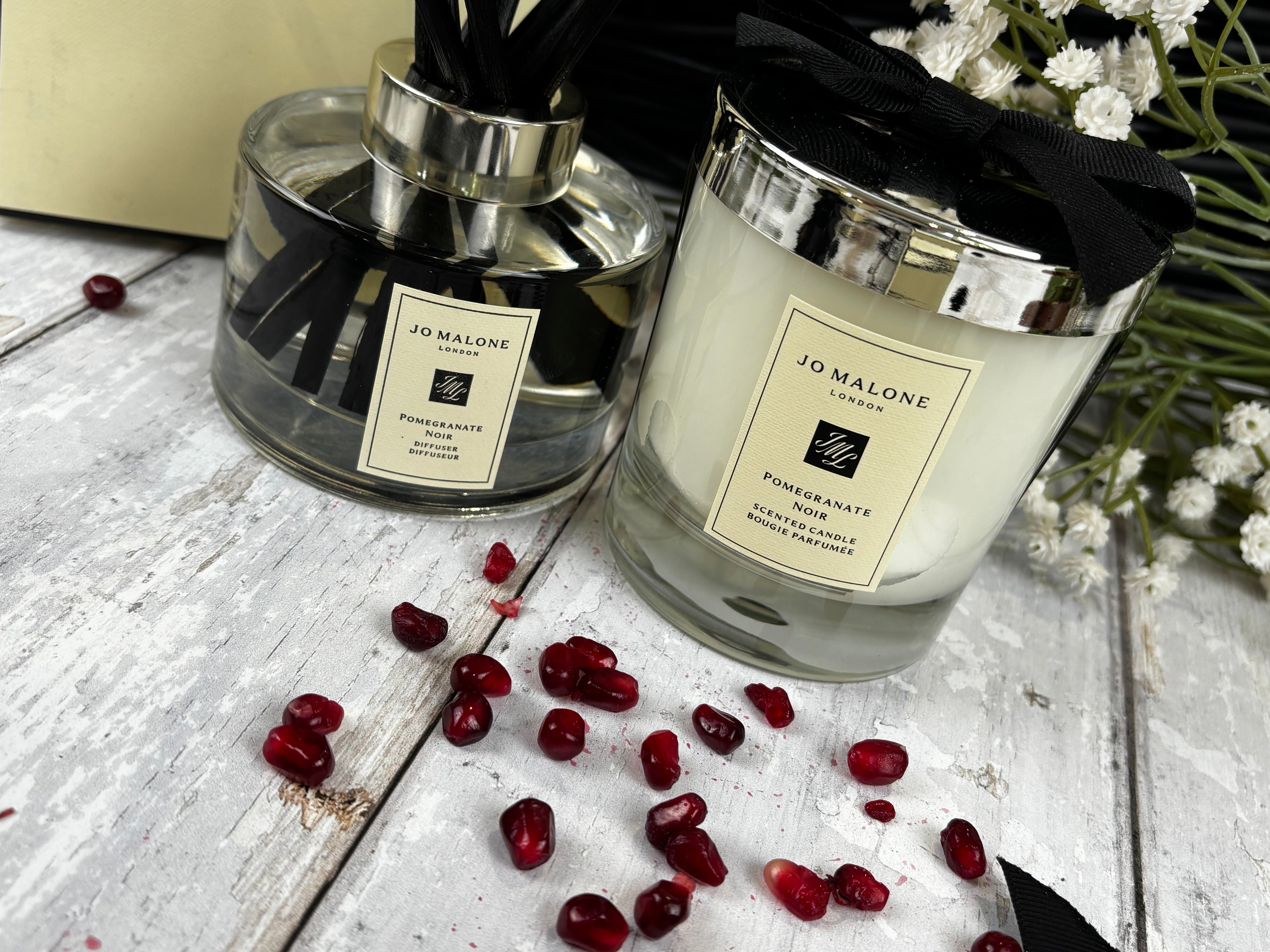 Jo Malone London Pomegranate Scented Candle and matching reed diffuser set on a rustic white wooden surface with scattered pomegranate seeds and white baby's breath flowers.