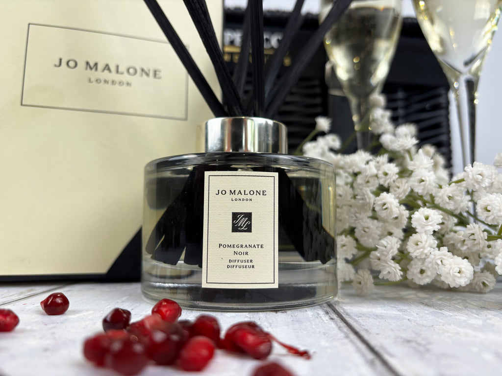 Jo Malone London Pomegranate Noir reed diffuser bottle with black reeds, set on a white surface with scattered pomegranate seeds, white flowers, and a branded box in the background.