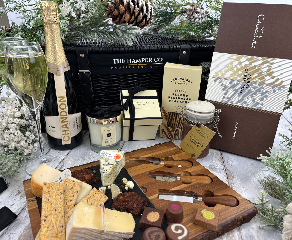 A festive gourmet gift hamper arrangement featuring a Chandon sparkling wine bottle, two filled champagne flutes, a black The Hamper Co gift bag, a Jo Malone candle, a box of crackers, a wooden charcuterie board with cheese and chocolates, and a Hotel Chocolat gift box, styled with pine branches and a pinecone on a white wooden surface.