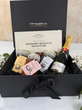 A luxury gift hamper from The Hamper Co featuring a Savoury Biscuits Selection by Cartwright & Butler, a bottle of Moët & Chandon, and small jars of preserves, all arranged inside an open black gift box with a black ribbon bow.