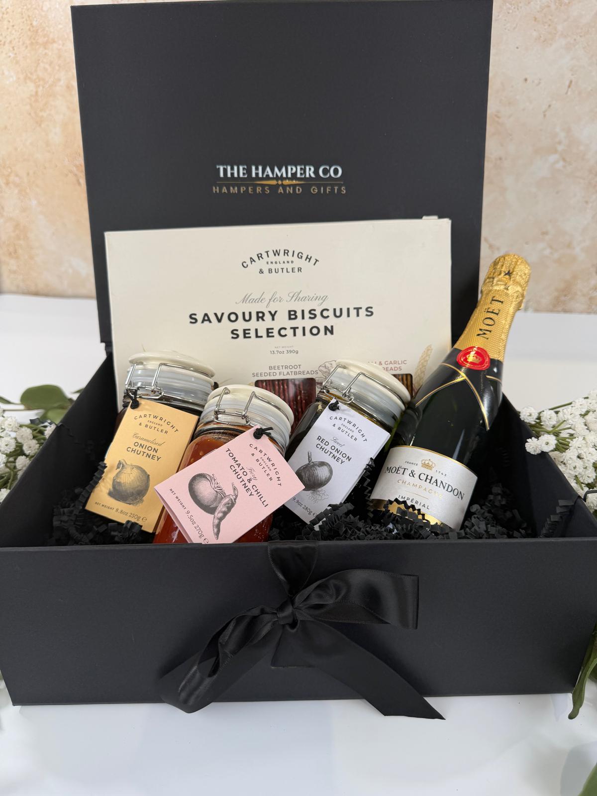 A luxury gift hamper from The Hamper Co featuring a Savoury Biscuits Selection by Cartwright & Butler, a bottle of Moët & Chandon, and small jars of preserves, all arranged inside an open black gift box with a black ribbon bow.