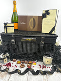 A luxury hamper gift set from The Hamper Co featuring a black wicker basket, a bottle of verve clicquot Champagne, chocolates, a  jo malone candle, and a jo malone reed diffuser, styled with black ribbons on a white wooden surface.