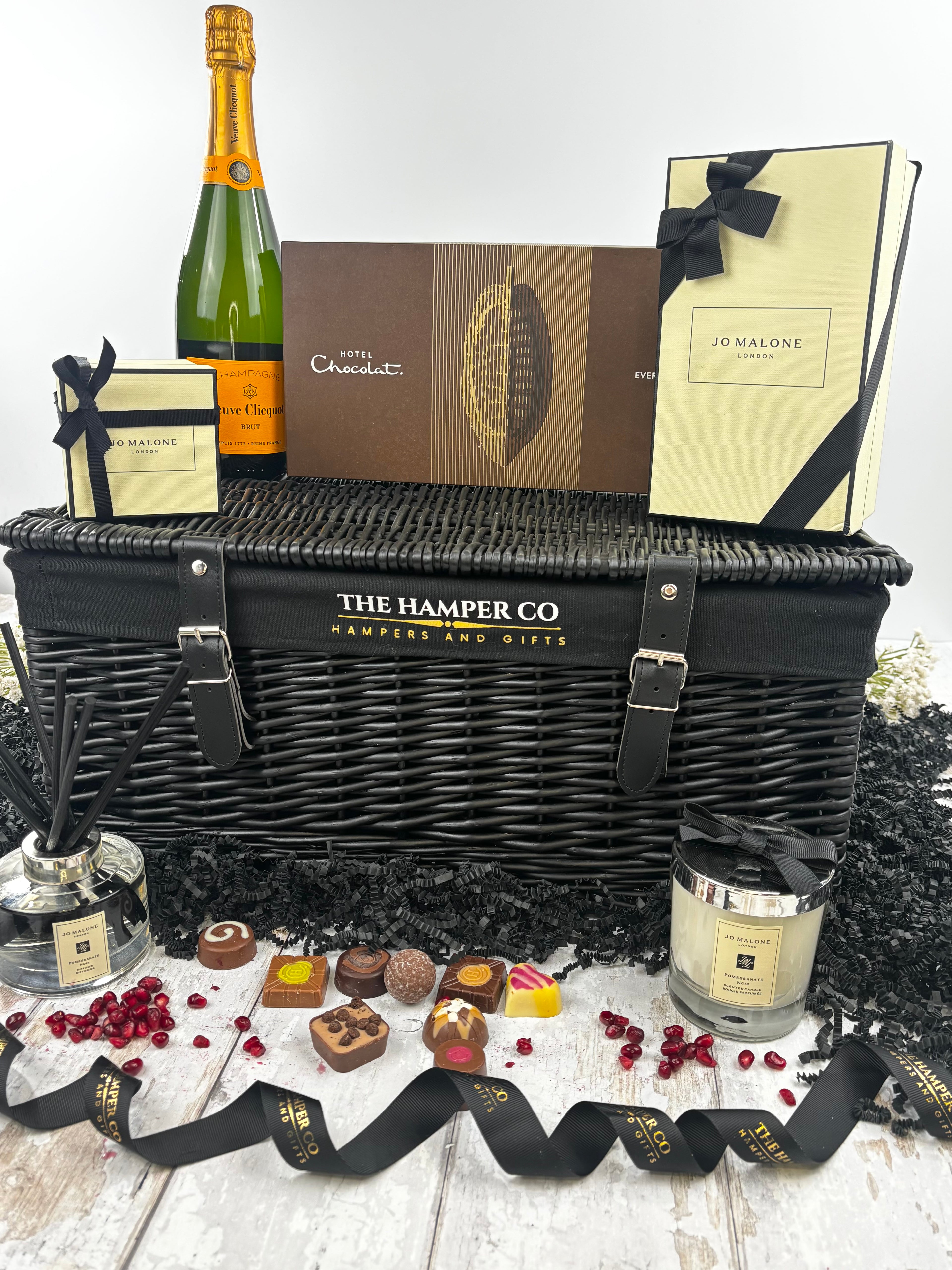 A luxury hamper gift set from The Hamper Co featuring a black wicker basket, a bottle of verve clicquot Champagne, chocolates, a  jo malone candle, and a jo malone reed diffuser, styled with black ribbons on a white wooden surface.