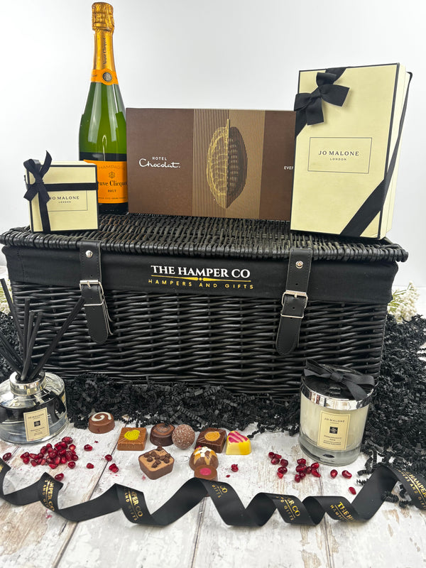 A luxury hamper gift set from The Hamper Co featuring a black wicker basket, a bottle of verve clicquot Champagne, chocolates, a  jo malone candle, and a jo malone reed diffuser, styled with black ribbons on a white wooden surface.