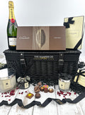 A luxury hamper gift set from The Hamper Co featuring a black wicker basket, a bottle of Moet Chardon champagne, Jo Malone candles, hotel chocolat chocolates, and gift boxes, arranged on a white wooden surface.