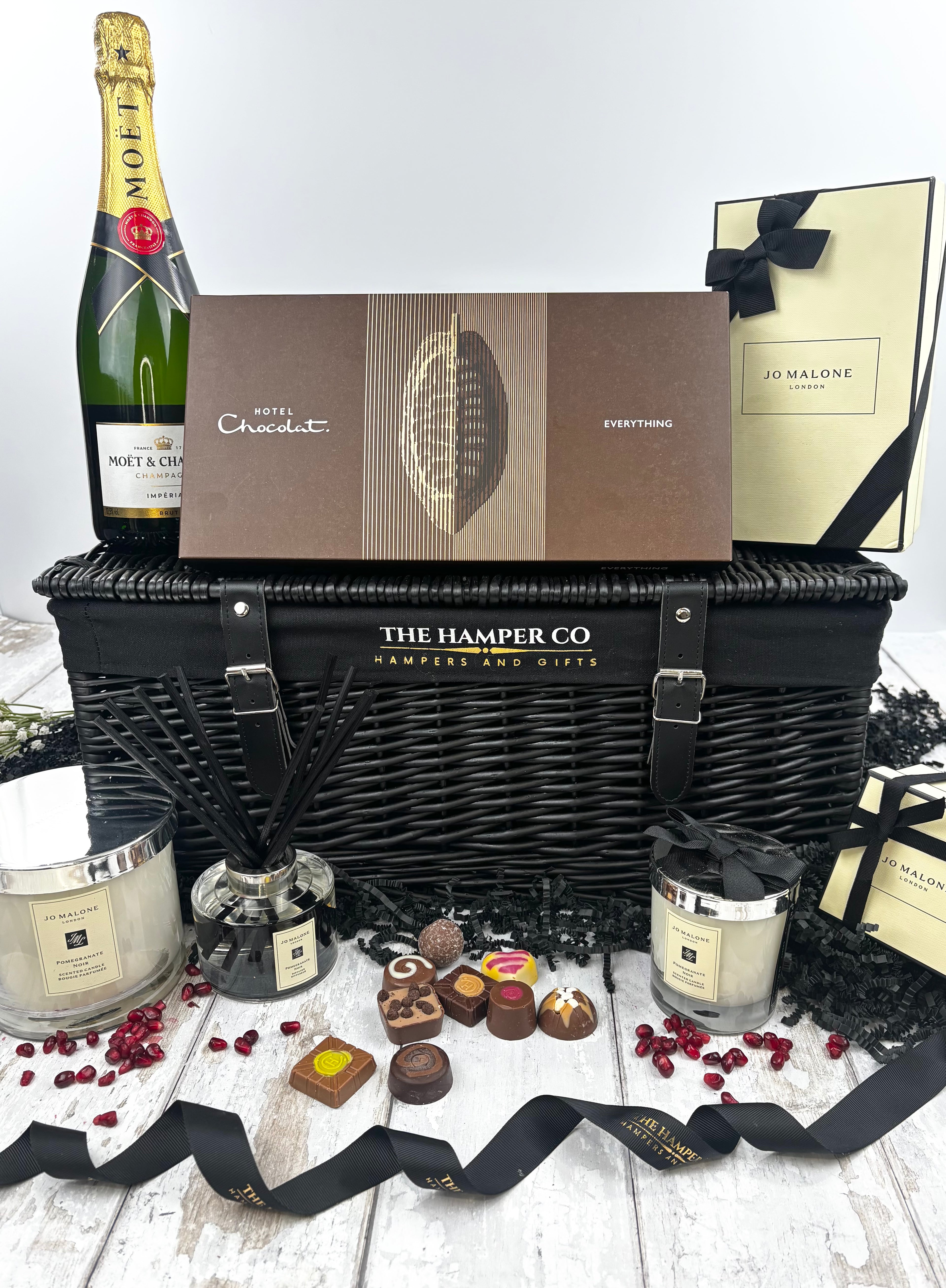 A luxury hamper gift set from The Hamper Co featuring a black wicker basket, a bottle of Moet Chardon champagne, Jo Malone candles, hotel chocolat chocolates, and gift boxes, arranged on a white wooden surface.