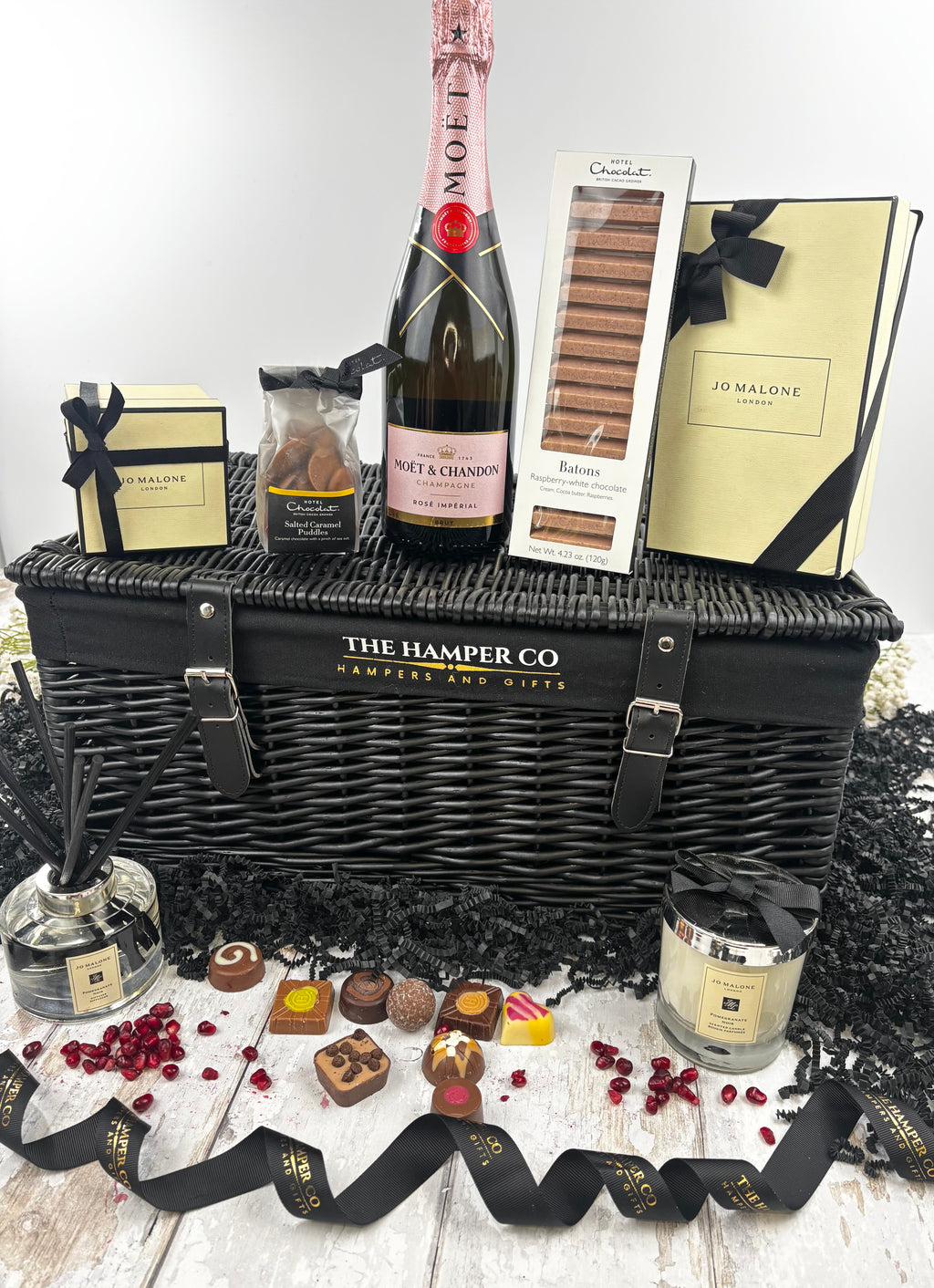 A luxury hamper gift set from The Hamper Co featuring a black wicker picnic basket with a Moët & Chardon sparkling wine bottle, hotel chocolat assorted chocolates, ajo malone  reed diffuser and candle. arranged on a white wood surface with black shredded paper.
