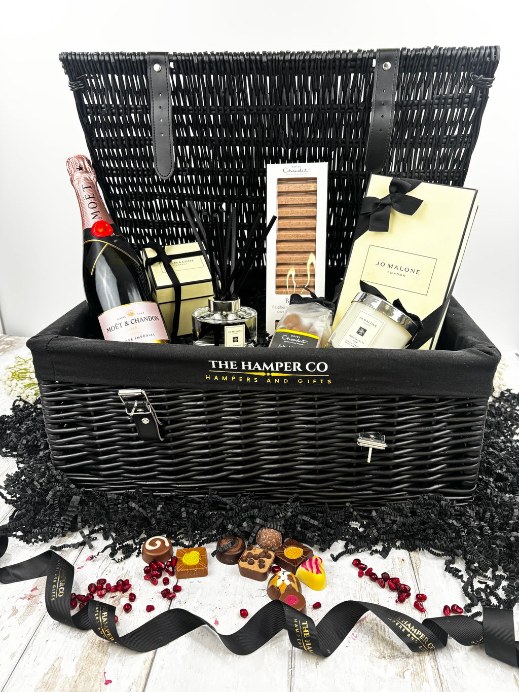 A luxury hamper gift set from The Hamper Co featuring a black wicker picnic basket with a black fabric liner, filled with a bottle of champagne, hotel chocolat, a jo malone  candle and diffuser,  displayed on a white wooden surface with decorative black paper shred and chocolates in front.