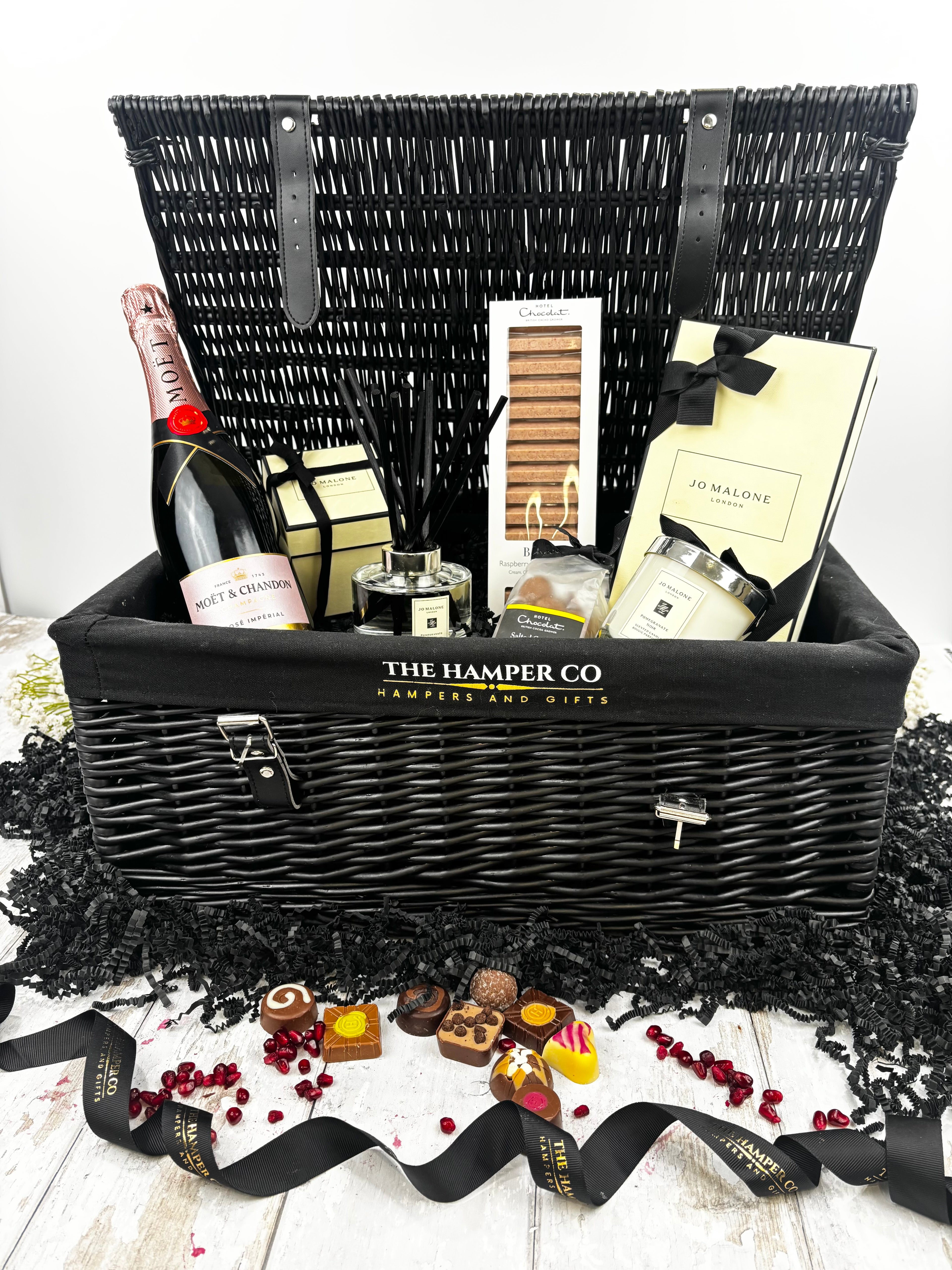 A luxury hamper gift set from The Hamper Co featuring a black wicker picnic basket with a black fabric liner, filled with a bottle of champagne, hotel chocolat, a jo malone  candle and diffuser,  displayed on a white wooden surface with decorative black paper shred and chocolates in front.