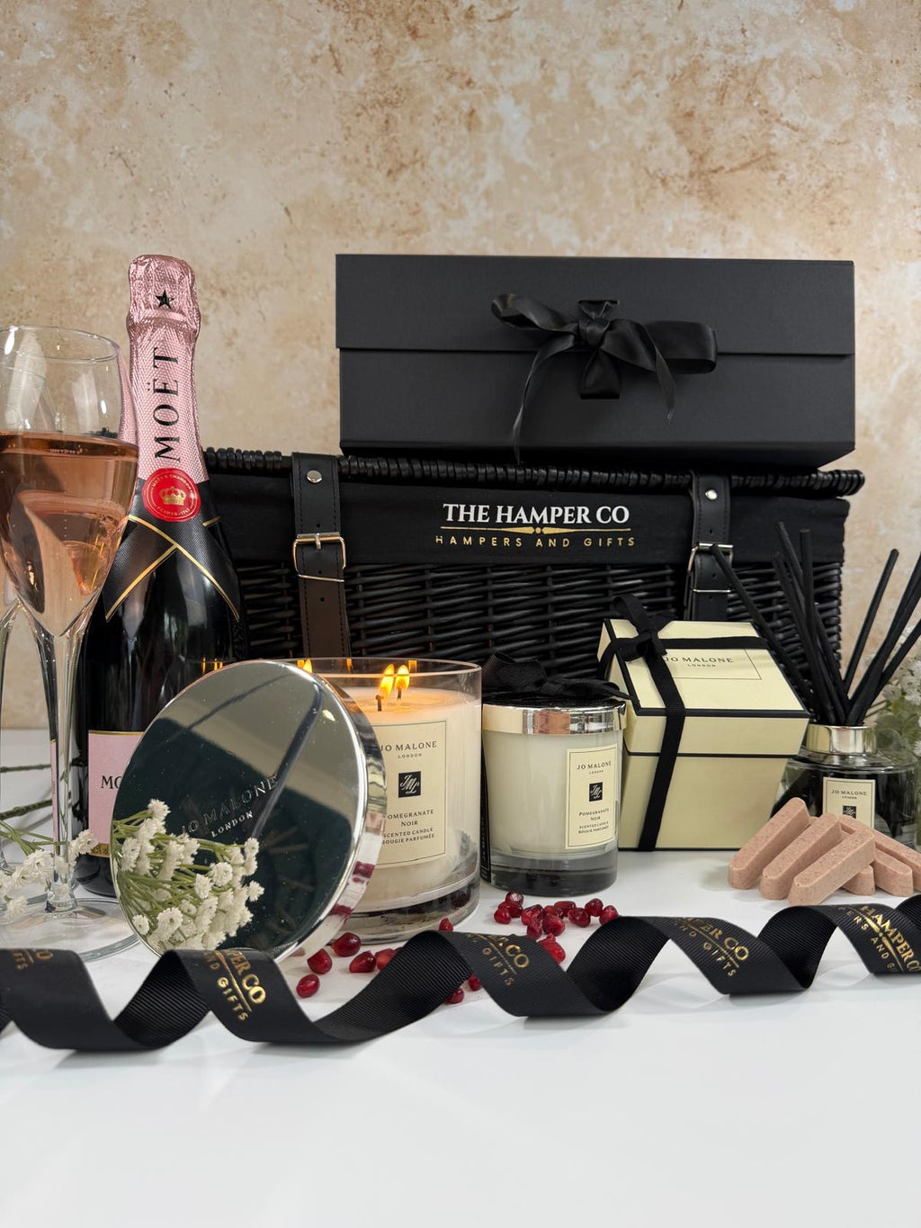 A luxury gift hamper from The Hamper Co arranged on a white surface against a textured beige wall. The set includes a black wicker basket with a black lid, a Moët champagne bottle, a lit jo malone candle & reed diffuser, and hotel chocolat pink finger chocolates, with black branded ribbons.