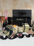 A luxury gift hamper from The Hamper Co arranged on a white surface against a textured beige wall. The set includes a black wicker basket with a black lid, a Moët champagne bottle, a lit jo malone candle & reed diffuser, and hotel chocolat pink finger chocolates, with black branded ribbons.