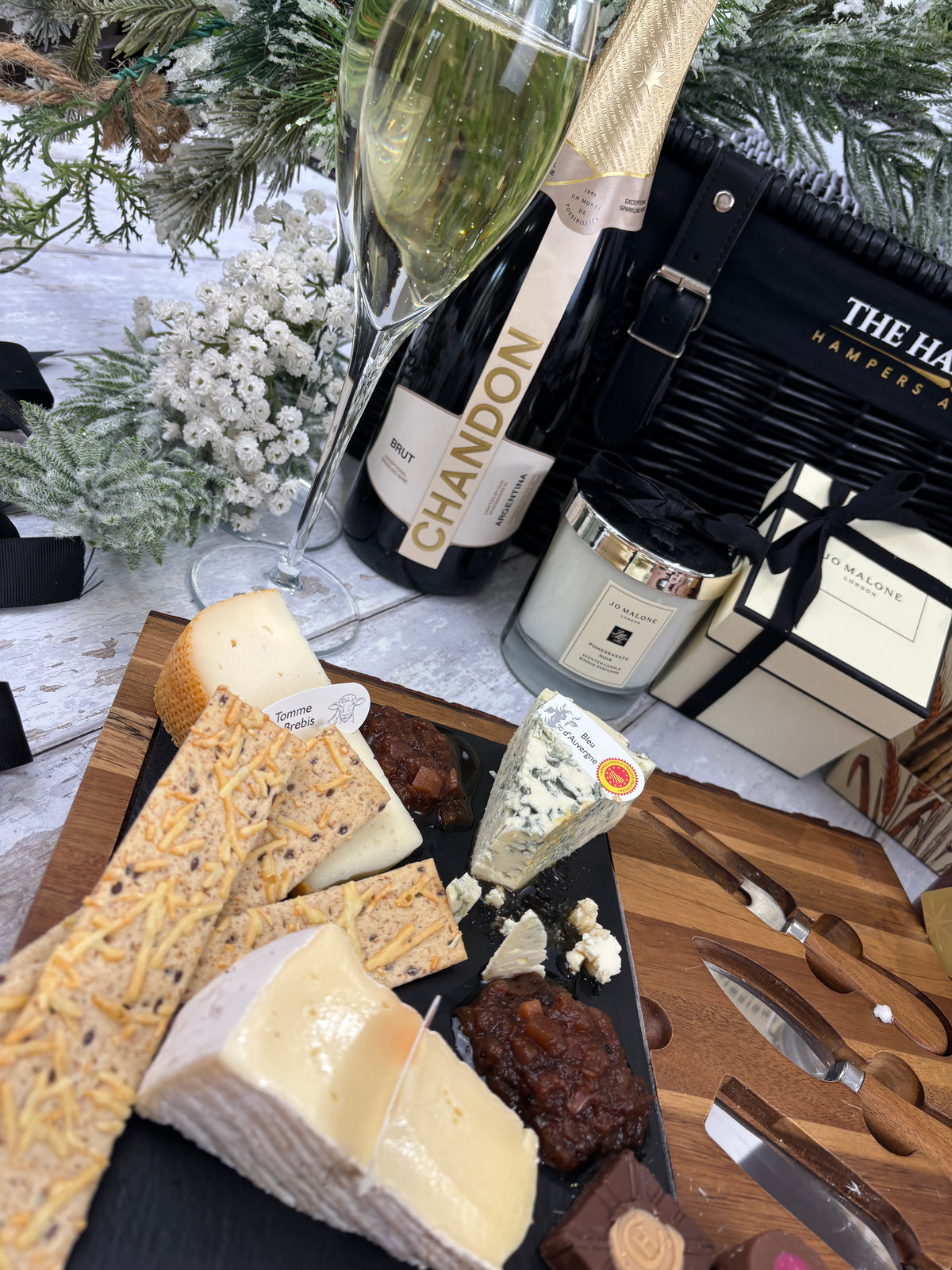 A styled charcuterie and cheese board with assorted cheeses, crackers, and cured meats on a wooden board, alongside a wine bottle, a filled wine glass, a candle, and a black gift basket from The Hamper Co on a white surface with holiday greenery in the background.