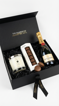 Gift box containing a bottle of champagne,  jo malone candle, and hotel chocolat macarons with 'The Hamper Co.' branding.