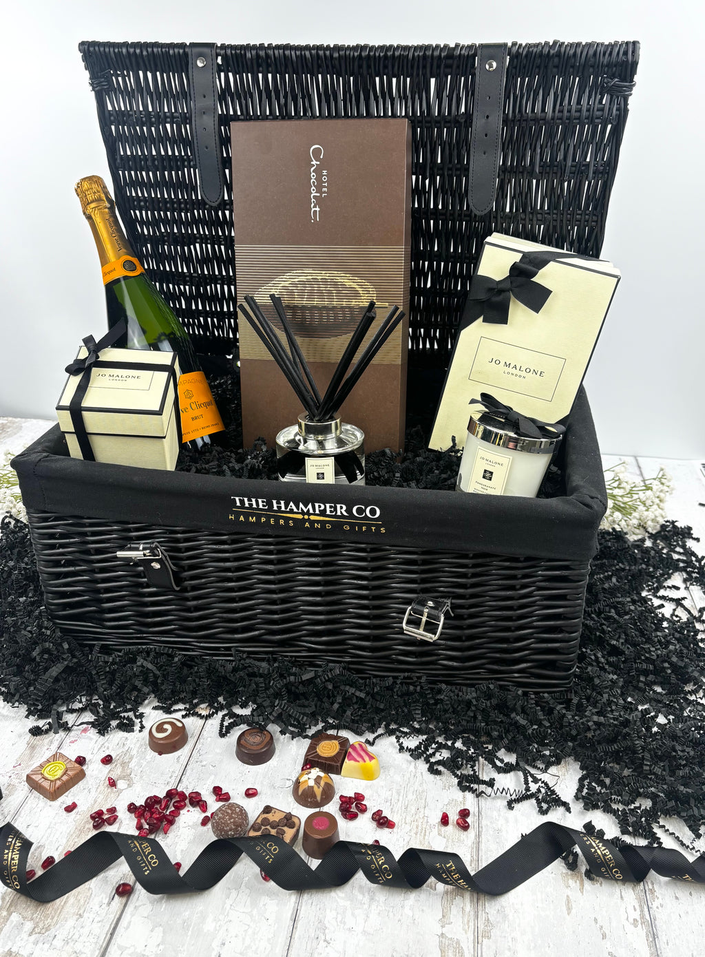 A luxury hamper gift set from The Hamper Co, featuring a black wicker picnic basket filled with a bottle of champagne, a reed diffuser, a candle, and a chocolate box, styled with scattered black paper shred, chocolates, and a black ribbon on a white wood background.