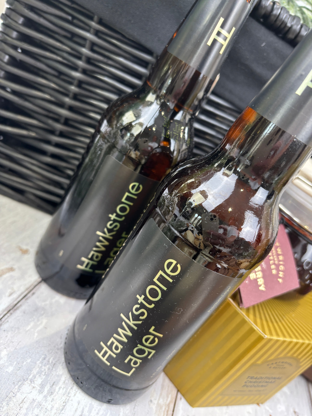Two dark glass bottles of Hawkstone Lager beer with black labels and gold lettering, set on a light wooden surface next to a gold box.